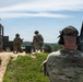 Military personnel are conducting a small arms range training exercise at an outdoor firing range. A soldier identified by name tape as 'ARDNER' is observing from a range control position, wearing tactical hearing protection. Multiple soldiers in OCP (Operational Camouflage Pattern) uniforms are visible downrange in various firing positions.