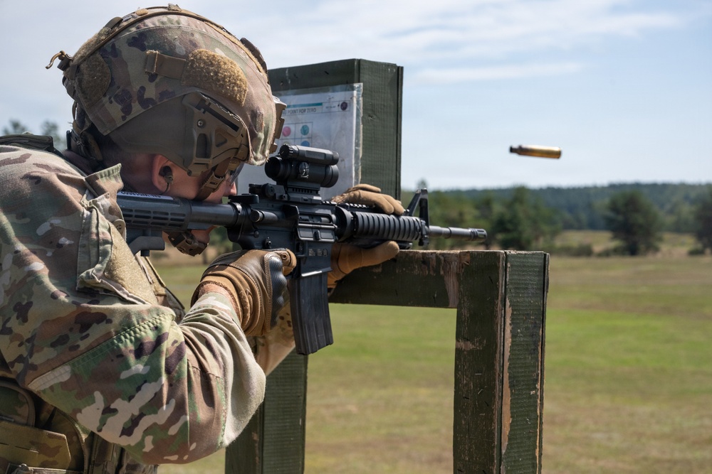 A soldier in full multicam combat gear is firing an M4/M16-style rifle at a military shooting range. A brass cartridge casing is visibly ejecting from the weapon mid-shot. The setting appears to be an outdoor zeroing or qualification range with a target board visible in the background.