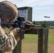 A soldier in full multicam combat gear is firing an M4/M16-style rifle at a military shooting range. A brass cartridge casing is visibly ejecting from the weapon mid-shot. The setting appears to be an outdoor zeroing or qualification range with a target board visible in the background.
