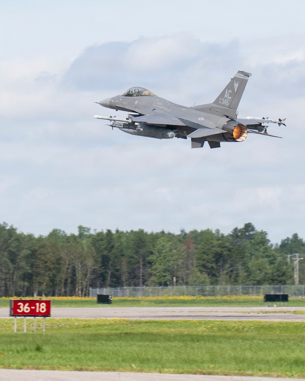 An F-16 Fighting Falcon jet fighter is captured just after takeoff from a military airfield, with its afterburner visibly glowing orange. The aircraft bears tail code 'AC' and serial number '345', consistent with the New Jersey Air National Guard's 177th Fighter Wing. A runway designation sign reading '36-18' is visible in the foreground, with a tree line in the background.