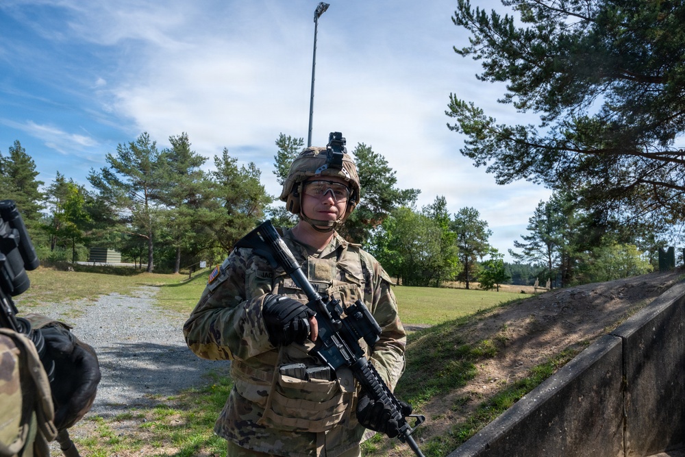 A U.S. Army soldier in full combat gear stands alert along a gravel path in what appears to be a training area or military installation. The soldier is equipped with an M4-style carbine, tactical helmet with accessories, and MultiCam/OCP camouflage uniform. The setting features pine trees, open grass fields, and a concrete barrier, consistent with a military training range environment.