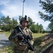 A U.S. Army soldier in full combat gear stands alert along a gravel path in what appears to be a training area or military installation. The soldier is equipped with an M4-style carbine, tactical helmet with accessories, and MultiCam/OCP camouflage uniform. The setting features pine trees, open grass fields, and a concrete barrier, consistent with a military training range environment.