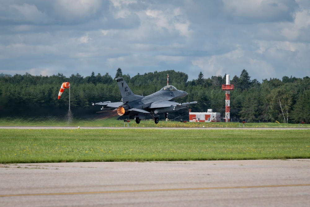 An F-16 Fighting Falcon jet fighter is captured during takeoff on a military airfield runway, with afterburner visibly ignited showing an orange flame at the rear engine. The aircraft bears 'AC' tail codes and the number '366' on the vertical stabilizer, indicating it belongs to the Atlantic City Air National Guard (177th Fighter Wing). The setting appears to be a well-maintained airfield surrounded by dense tree lines under a partly cloudy sky.
