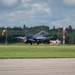An F-16 Fighting Falcon jet fighter is captured during takeoff on a military airfield runway, with afterburner visibly ignited showing an orange flame at the rear engine. The aircraft bears 'AC' tail codes and the number '366' on the vertical stabilizer, indicating it belongs to the Atlantic City Air National Guard (177th Fighter Wing). The setting appears to be a well-maintained airfield surrounded by dense tree lines under a partly cloudy sky.