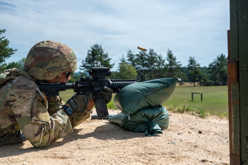 A soldier in full camouflage uniform and combat helmet is firing an M4/M16-style rifle from a prone or low-kneeling position at a military shooting range. A brass cartridge casing is visibly ejecting from the weapon, captured mid-air. The soldier is using sandbags as a rest or barrier, with a downrange field and target frames visible in the background.