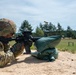 A soldier in full camouflage uniform and combat helmet is firing an M4/M16-style rifle from a prone or low-kneeling position at a military shooting range. A brass cartridge casing is visibly ejecting from the weapon, captured mid-air. The soldier is using sandbags as a rest or barrier, with a downrange field and target frames visible in the background.
