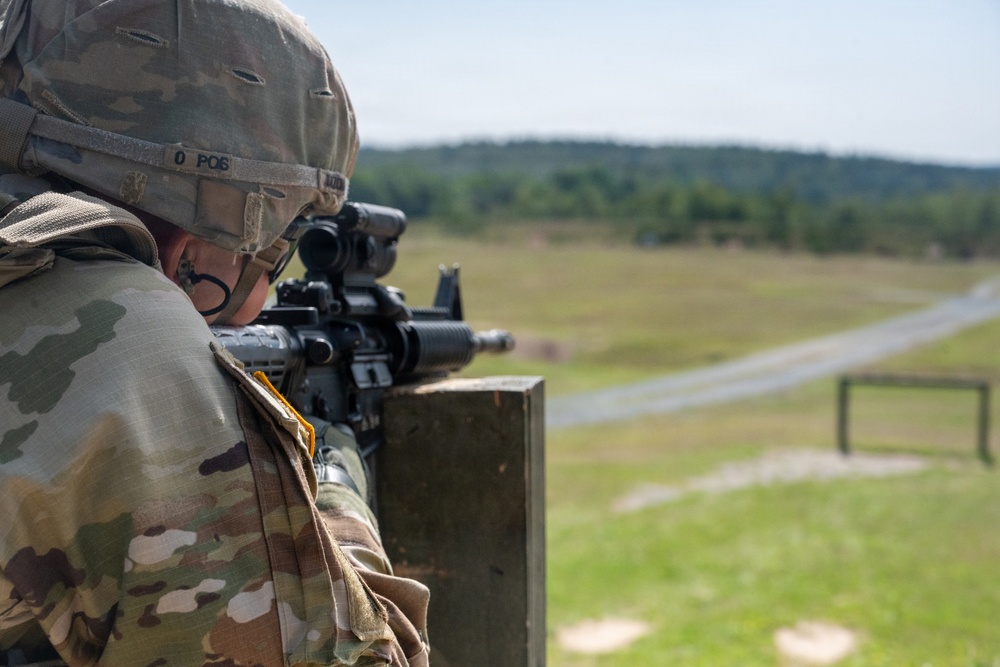 A soldier in MultiCam/OCP camouflage uniform is in a prone or supported firing position at what appears to be a military rifle range. The soldier is aiming an M4/M16-style carbine equipped with an optical sight over a wooden barricade. The background shows a typical military training range with open fields, a road, and treeline.