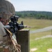 A soldier in MultiCam/OCP camouflage uniform is in a prone or supported firing position at what appears to be a military rifle range. The soldier is aiming an M4/M16-style carbine equipped with an optical sight over a wooden barricade. The background shows a typical military training range with open fields, a road, and treeline.
