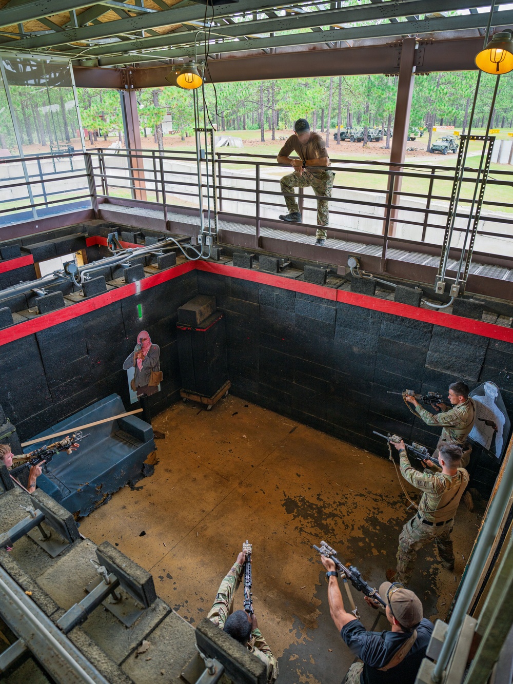 An overhead view of a military or law enforcement close-quarters battle (CQB) training exercise taking place inside an indoor shoot house facility. Multiple personnel in tactical/military uniforms are positioned around a room with weapons raised, engaging simulated targets including a cardboard cutout of a hostile figure. One individual is visible on an elevated observation catwalk above the training area.