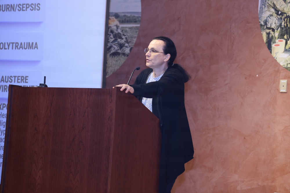 A woman is presenting at a podium during what appears to be a medical or military medicine conference. She is gesturing toward a projection screen displaying topics including 'BURN/SEPSIS', 'POLYTRAUMA', and 'AUSTERE ENVIRONMENT'. The setting appears to be a formal conference room with a terracotta-colored wall and military-themed imagery in the background.