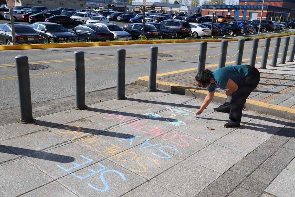 A person wearing a teal/green top is bent over writing chalk art on a paved sidewalk near a parking area. The chalk writing appears to read 'SAY SOMETHING' in large, colorful multicolored letters. The scene is set in what appears to be a commercial or public area with bollards, a yellow chain barrier, and a large parking lot visible in the background.