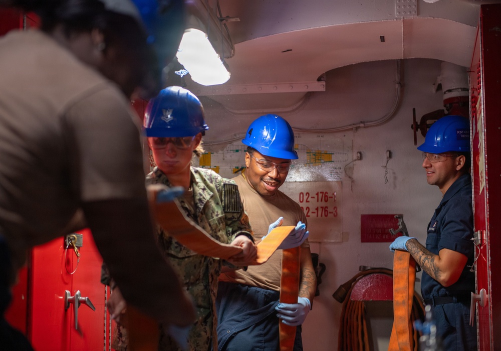 Several military personnel wearing blue hard hats and safety glasses are working inside what appears to be a naval vessel compartment. They appear to be conducting an inspection or inventory task, with at least one person reviewing documents or materials. The group includes personnel in Navy camouflage uniforms and Coast Guard-style dark blue uniforms.