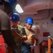 Several military personnel wearing blue hard hats and safety glasses are working inside what appears to be a naval vessel compartment. They appear to be conducting an inspection or inventory task, with at least one person reviewing documents or materials. The group includes personnel in Navy camouflage uniforms and Coast Guard-style dark blue uniforms.