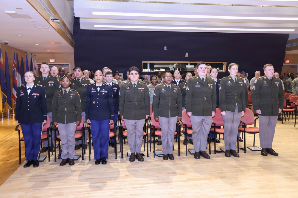 A formal military ceremony is taking place indoors, with a row of uniformed U.S. Army soldiers standing at attention in the foreground. The soldiers are wearing Army Service Uniforms (ASUs) in green and blue variants, with visible rank insignia and unit patches. A larger group of uniformed personnel is visible in the background, along with military flags on the left side.