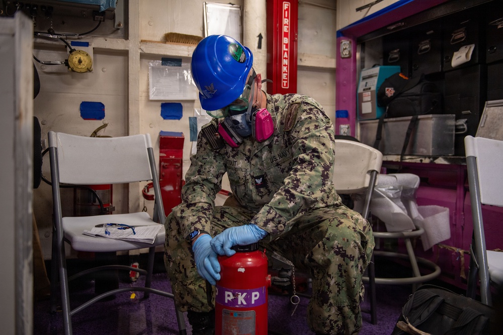 A U.S. Navy sailor wearing personal protective equipment (blue hard hat, respirator mask, safety goggles, and blue nitrile gloves) is inspecting or servicing a red PKP (Purple-K Powder) fire extinguisher in what appears to be an interior compartment of a naval vessel. The sailor is dressed in Navy Working Uniform (NWU) camouflage pattern with visible rank insignia and a name tape reading 'STOUT'. The setting includes fire safety equipment and storage areas consistent with a shipboard environment.