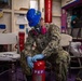 A U.S. Navy sailor wearing personal protective equipment (blue hard hat, respirator mask, safety goggles, and blue nitrile gloves) is inspecting or servicing a red PKP (Purple-K Powder) fire extinguisher in what appears to be an interior compartment of a naval vessel. The sailor is dressed in Navy Working Uniform (NWU) camouflage pattern with visible rank insignia and a name tape reading 'STOUT'. The setting includes fire safety equipment and storage areas consistent with a shipboard environment.