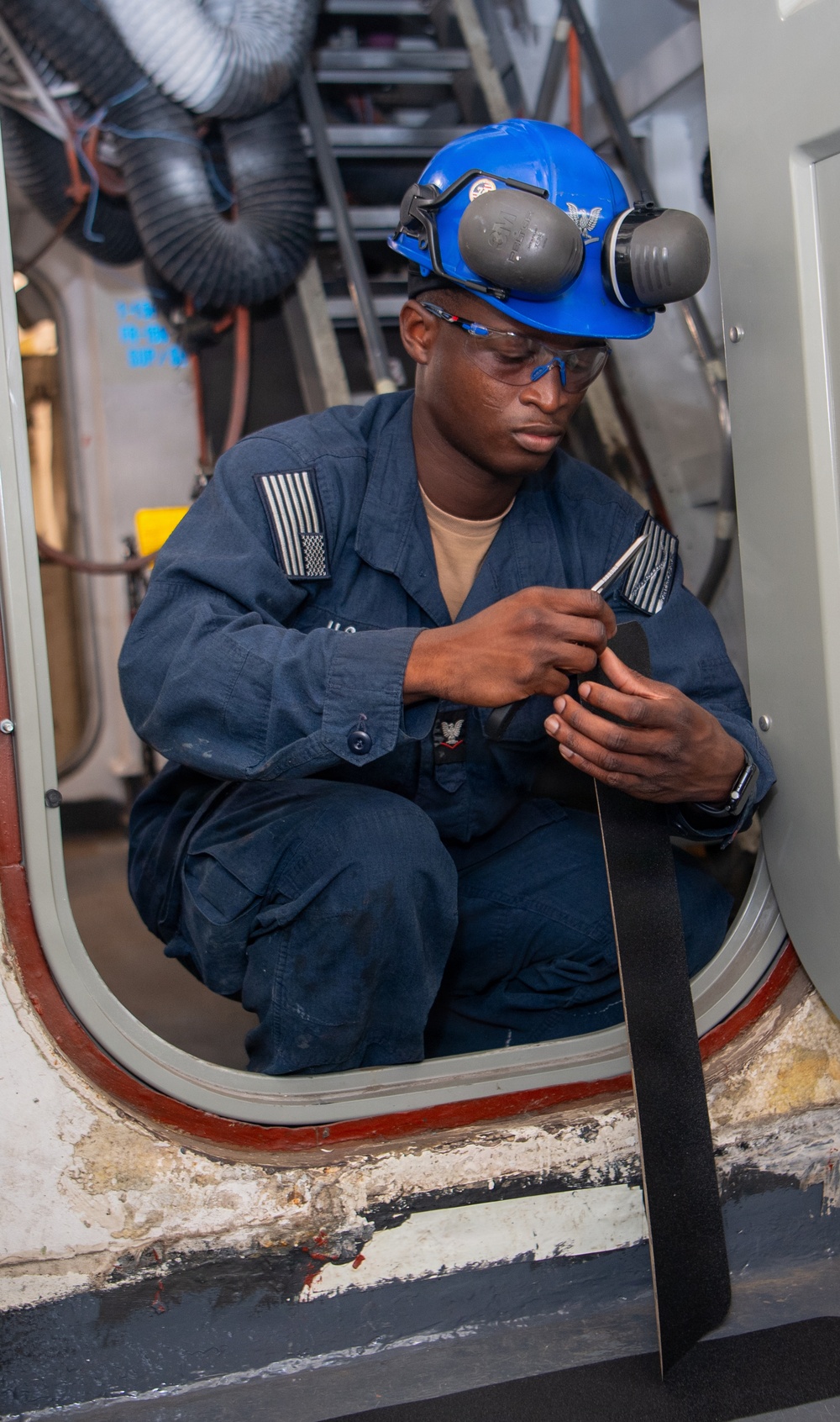 A U.S. Navy sailor in blue coveralls is crouched in a watertight hatch/door opening aboard a naval vessel, working with a tool or component in his hands. He is wearing full personal protective equipment including a blue hard hat, ear protection muffs, and safety glasses. The setting appears to be a shipboard maintenance or repair environment.