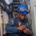 A U.S. Navy sailor in blue coveralls is crouched in a watertight hatch/door opening aboard a naval vessel, working with a tool or component in his hands. He is wearing full personal protective equipment including a blue hard hat, ear protection muffs, and safety glasses. The setting appears to be a shipboard maintenance or repair environment.