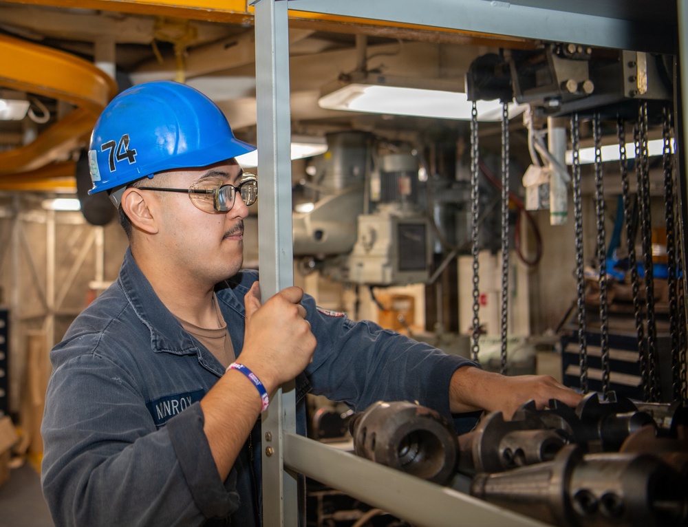 A worker wearing a blue hard hat marked with the number '74' and glasses is working in an industrial or shipboard machinery space. The individual, whose name tag reads 'NNROX', is handling mechanical components on a metal shelving rack surrounded by chains and heavy equipment. The setting appears to be a maintenance or machine shop environment, likely aboard a naval vessel.