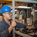 A worker wearing a blue hard hat marked with the number '74' and glasses is working in an industrial or shipboard machinery space. The individual, whose name tag reads 'NNROX', is handling mechanical components on a metal shelving rack surrounded by chains and heavy equipment. The setting appears to be a maintenance or machine shop environment, likely aboard a naval vessel.