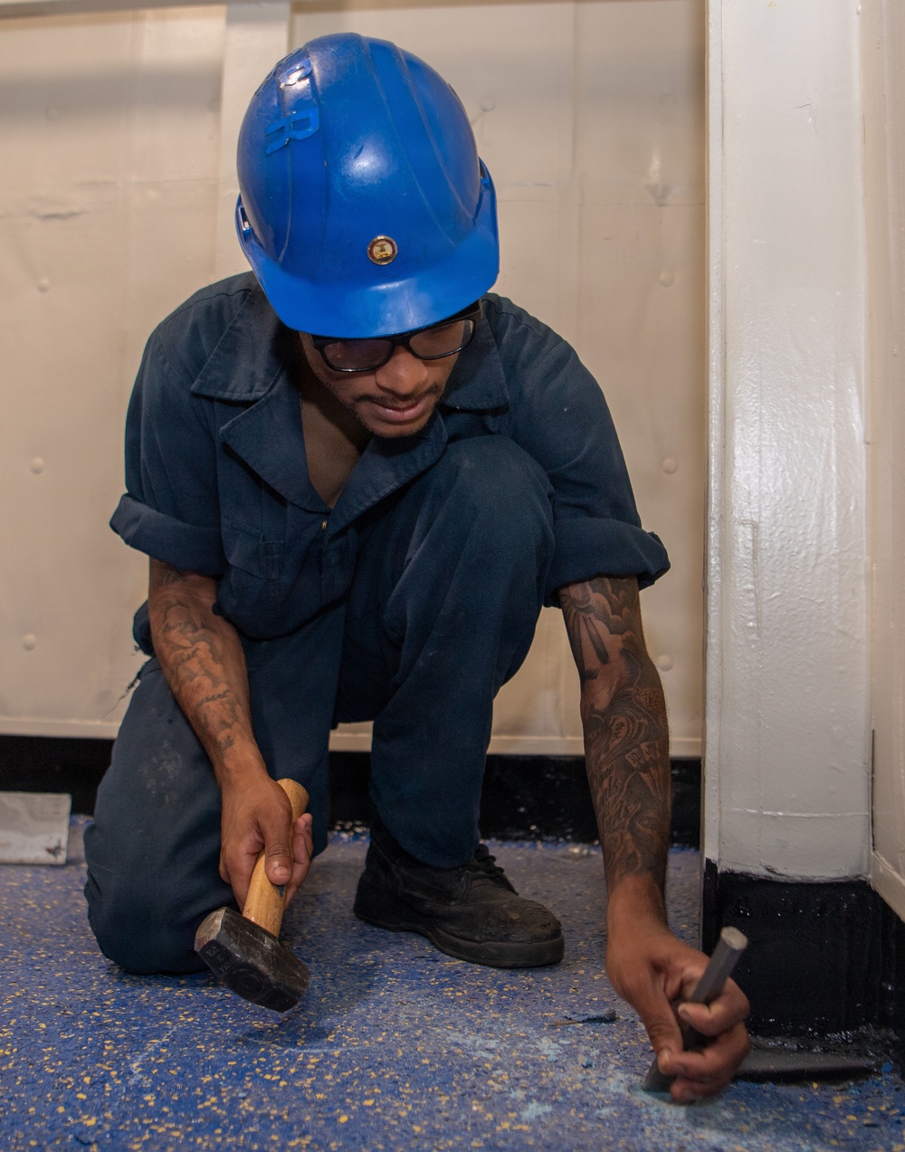 A worker wearing a blue hard hat, safety glasses, and dark navy coveralls is kneeling on a blue speckled floor performing maintenance work. He is using a hammer and chisel or punch tool near the base of a white painted wall or structural column. The worker has visible tattoos on both forearms and appears focused on the task at hand.