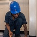 A worker wearing a blue hard hat, safety glasses, and dark navy coveralls is kneeling on a blue speckled floor performing maintenance work. He is using a hammer and chisel or punch tool near the base of a white painted wall or structural column. The worker has visible tattoos on both forearms and appears focused on the task at hand.