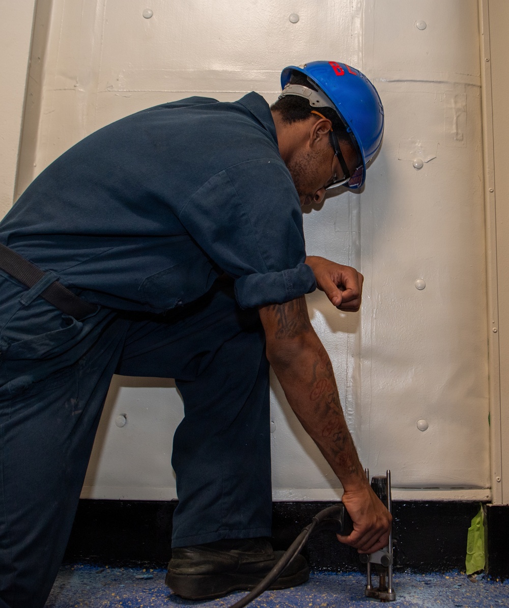 A worker wearing a blue hard hat and navy blue coveralls is bent over performing maintenance or repair work at the base of a white metal wall or bulkhead. He is using a power tool at floor level, suggesting drilling, grinding, or fastening work. The setting appears to be an industrial or maritime environment based on the painted metal surfaces and floor.