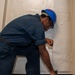 A worker wearing a blue hard hat and navy blue coveralls is bent over performing maintenance or repair work at the base of a white metal wall or bulkhead. He is using a power tool at floor level, suggesting drilling, grinding, or fastening work. The setting appears to be an industrial or maritime environment based on the painted metal surfaces and floor.