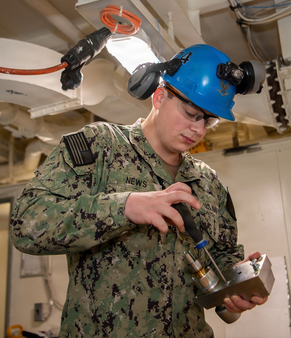 A U.S. Navy sailor wearing NWU Type III camouflage uniform is performing a technical task in what appears to be a shipboard interior space. The sailor, identified by name tape as 'NEWS,' is wearing a blue hard hat marked '74,' safety glasses, ear protection, and a headlamp while handling a tool or instrument connected to a metal component. The setting appears to be a maintenance or inspection activity aboard a naval vessel.