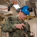 A U.S. Navy sailor wearing NWU Type III camouflage uniform is performing a technical task in what appears to be a shipboard interior space. The sailor, identified by name tape as 'NEWS,' is wearing a blue hard hat marked '74,' safety glasses, ear protection, and a headlamp while handling a tool or instrument connected to a metal component. The setting appears to be a maintenance or inspection activity aboard a naval vessel.