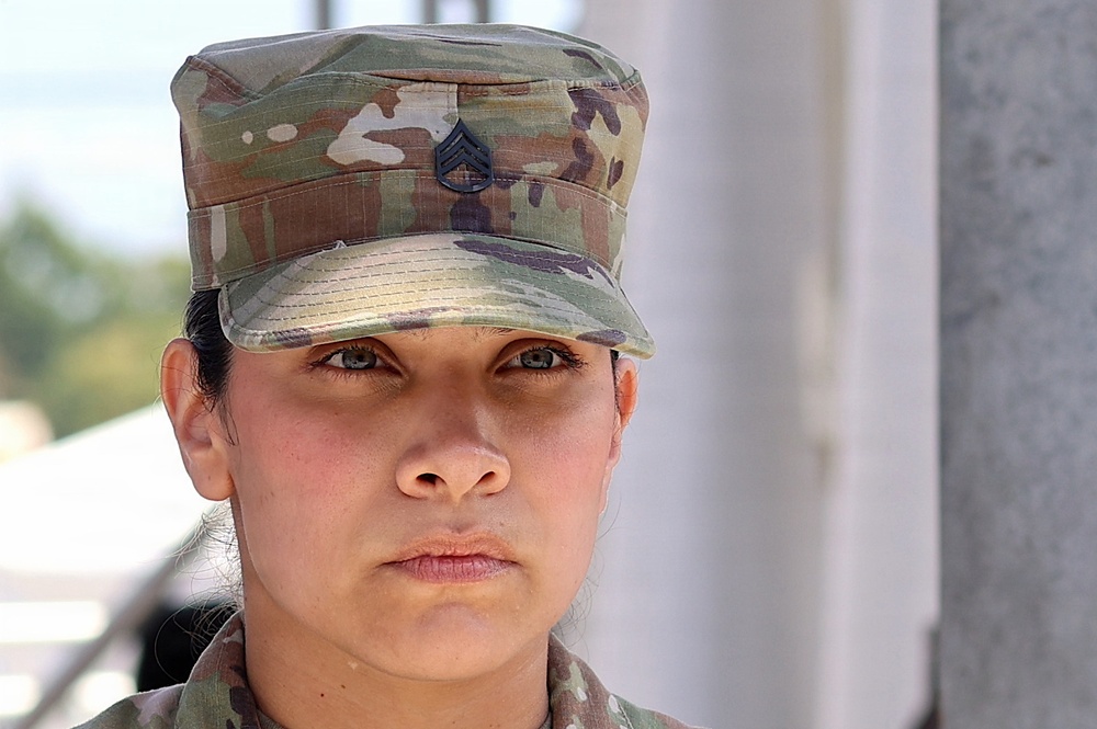 A close-up portrait of a female U.S. Army soldier wearing Operational Camouflage Pattern (OCP) uniform and patrol cap. A Staff Sergeant rank insignia is visible on the front of her cap. She has a serious, composed expression and is photographed against a blurred outdoor background.