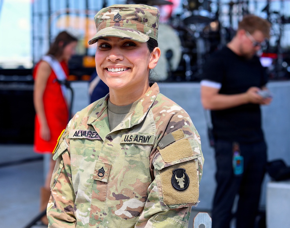 A U.S. Army soldier identified by name tape as 'Alvarez' poses smiling at what appears to be an outdoor event or concert venue. She wears the Operational Camouflage Pattern (OCP) uniform with Staff Sergeant rank insignia on her cap and chest, and a unit patch on her left sleeve. The background shows a stage setup with equipment and several civilians.