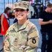 A U.S. Army soldier identified by name tape as 'Alvarez' poses smiling at what appears to be an outdoor event or concert venue. She wears the Operational Camouflage Pattern (OCP) uniform with Staff Sergeant rank insignia on her cap and chest, and a unit patch on her left sleeve. The background shows a stage setup with equipment and several civilians.