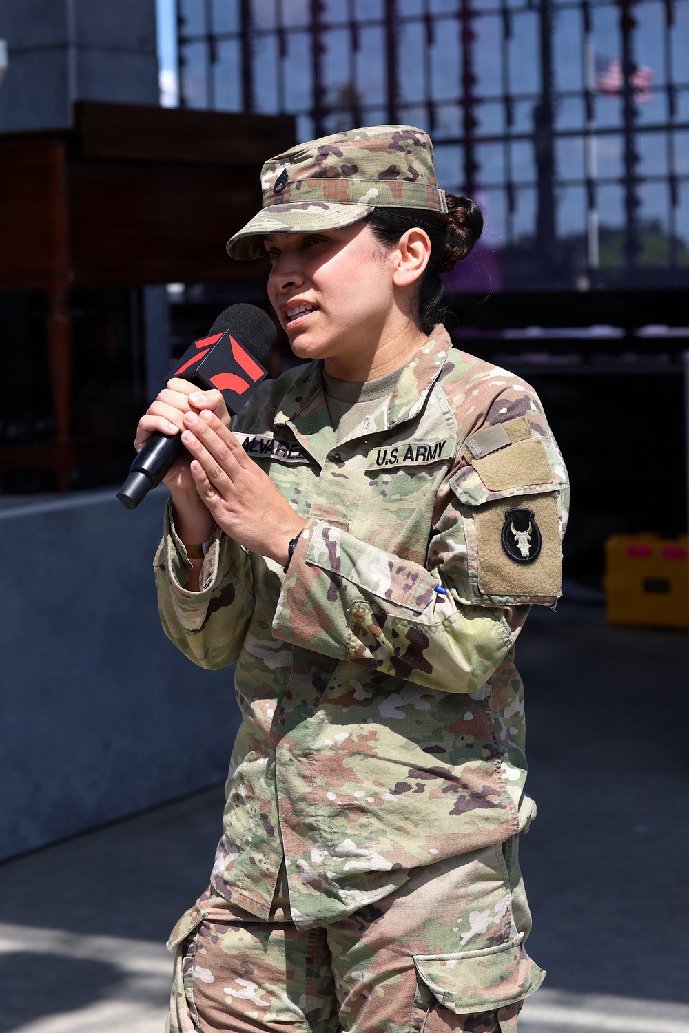 A female U.S. Army soldier in Operational Camouflage Pattern (OCP) uniform is speaking into a microphone at an outdoor event. Her name tape partially reads 'ALVAREZ' and she wears a unit patch on her left sleeve depicting a buffalo/bison skull. She appears to be giving a speech or interview in a public or ceremonial setting.