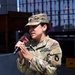 A female U.S. Army soldier in Operational Camouflage Pattern (OCP) uniform is speaking into a microphone at an outdoor event. Her name tape partially reads 'ALVAREZ' and she wears a unit patch on her left sleeve depicting a buffalo/bison skull. She appears to be giving a speech or interview in a public or ceremonial setting.