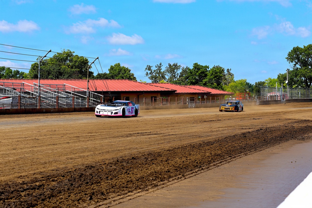 Two stock cars are racing on a dirt oval track at what appears to be a small regional racing facility. The foreground car is white with pink accents and bears the number 67, while the second car is yellow and blue. A red-roofed building and metal grandstands are visible in the background under a partly cloudy blue sky.