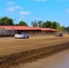 Two stock cars are racing on a dirt oval track at what appears to be a small regional racing facility. The foreground car is white with pink accents and bears the number 67, while the second car is yellow and blue. A red-roofed building and metal grandstands are visible in the background under a partly cloudy blue sky.