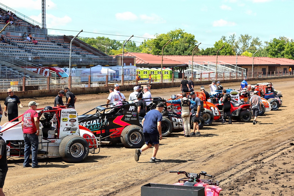 A dirt track racing event showing multiple sprint/modified race cars lined up in the pit or staging area on a dirt oval track. Crew members, drivers, and officials are gathered around the vehicles, suggesting pre-race preparations or a pit stop period. Grandstands with spectators are visible in the background along with facility buildings and portable toilets.