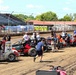 A dirt track racing event showing multiple sprint/modified race cars lined up in the pit or staging area on a dirt oval track. Crew members, drivers, and officials are gathered around the vehicles, suggesting pre-race preparations or a pit stop period. Grandstands with spectators are visible in the background along with facility buildings and portable toilets.