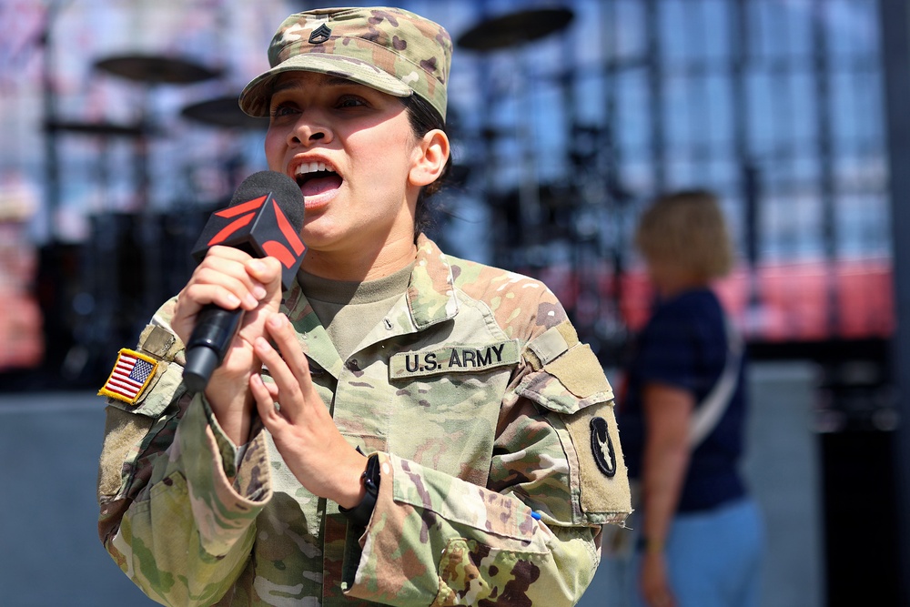 A female U.S. Army soldier in OCP (Operational Camouflage Pattern) uniform is singing into a microphone at what appears to be a public event or sporting venue. She holds a branded microphone and wears the rank of Staff Sergeant on her cap, with a U.S. flag patch and unit patch visible on her uniform. The background shows stadium seating and a stage setup, suggesting a pre-event ceremony or national anthem performance.