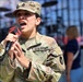 A female U.S. Army soldier in OCP (Operational Camouflage Pattern) uniform is singing into a microphone at what appears to be a public event or sporting venue. She holds a branded microphone and wears the rank of Staff Sergeant on her cap, with a U.S. flag patch and unit patch visible on her uniform. The background shows stadium seating and a stage setup, suggesting a pre-event ceremony or national anthem performance.