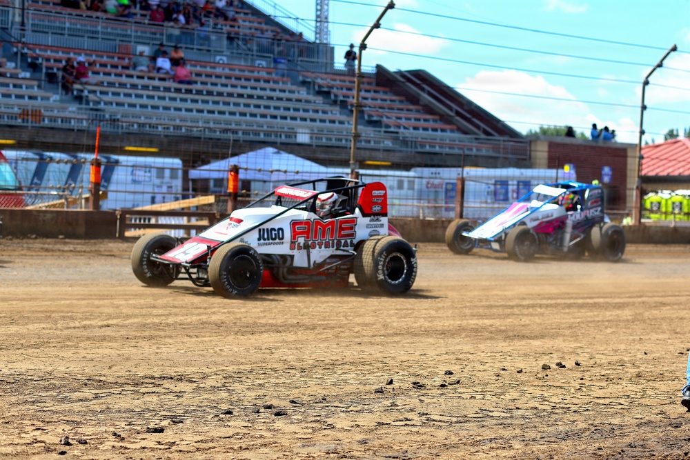 Two open-wheel midget or sprint cars are racing on a dirt track at what appears to be a fairgrounds or short oval circuit. The foreground car is prominently sponsored by 'AME Electrical' and 'JUGO Superfoods' in a white and red livery. Spectators are visible in the grandstands in the background under a partly cloudy sky.