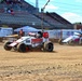 Two open-wheel midget or sprint cars are racing on a dirt track at what appears to be a fairgrounds or short oval circuit. The foreground car is prominently sponsored by 'AME Electrical' and 'JUGO Superfoods' in a white and red livery. Spectators are visible in the grandstands in the background under a partly cloudy sky.