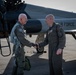 Two bald military aviators in olive/sage green flight suits are shaking hands on a flight line in front of a U.S. Air Force jet aircraft. The setting appears to be an outdoor tarmac during daylight hours, suggesting a formal or ceremonial greeting, possibly post-flight or a distinguished visitor event. The aircraft behind them bears 'AIR FORCE' lettering and a U.S. military star insignia.