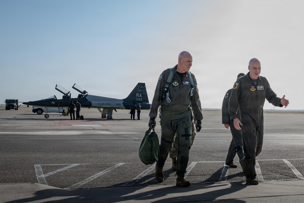 Two U.S. Air Force personnel in flight suits walk across a tarmac after what appears to be a flight mission. In the background, a T-38 Talon jet trainer aircraft marked 'RA 5143' and 'U.S. AIR FORCE' is parked on the flight line with ground crew nearby. The scene appears to be at a U.S. Air Force base, likely Randolph Air Force Base based on the 'RA' tail code.