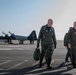 Two U.S. Air Force personnel in flight suits walk across a tarmac after what appears to be a flight mission. In the background, a T-38 Talon jet trainer aircraft marked 'RA 5143' and 'U.S. AIR FORCE' is parked on the flight line with ground crew nearby. The scene appears to be at a U.S. Air Force base, likely Randolph Air Force Base based on the 'RA' tail code.