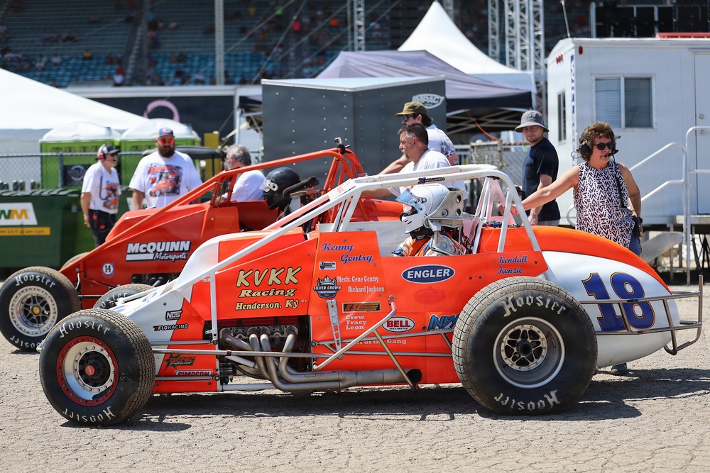 The image shows a racing paddock or pit area featuring two orange sprint/midget-style race cars, with car number 18 prominently in the foreground bearing 'KVKK Racing, Henderson, KY' branding. A helmeted driver is seated in car 18, while crew members, officials, and spectators mill about in the background. The setting appears to be a major racing event, likely at a large oval track facility.