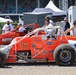 The image shows a racing paddock or pit area featuring two orange sprint/midget-style race cars, with car number 18 prominently in the foreground bearing 'KVKK Racing, Henderson, KY' branding. A helmeted driver is seated in car 18, while crew members, officials, and spectators mill about in the background. The setting appears to be a major racing event, likely at a large oval track facility.