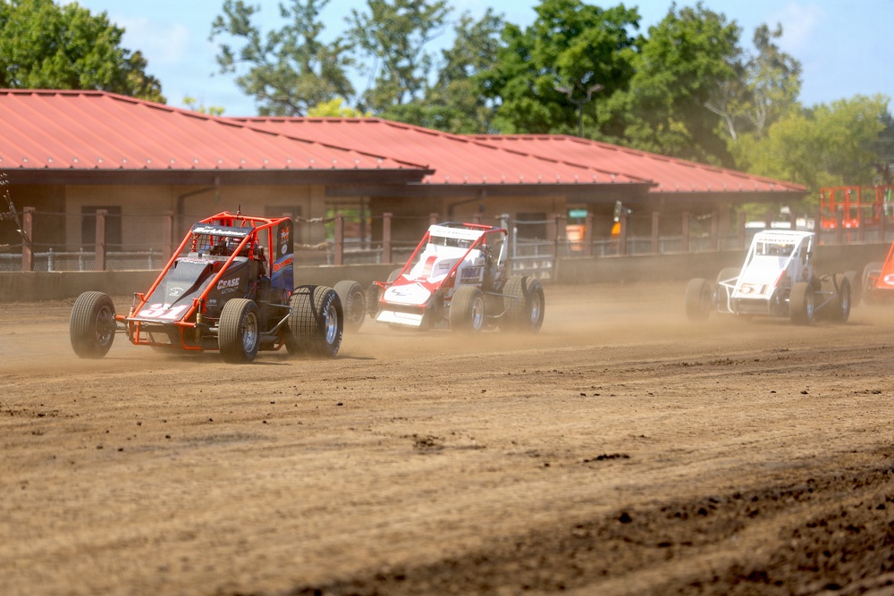 A dirt track racing event featuring multiple open-wheel sprint cars or midget race cars competing on a dusty oval circuit. At least three vehicles are visible racing in close proximity, kicking up significant dust clouds. The race appears to be taking place at a fairgrounds-style venue with a red-roofed building and trees visible in the background.