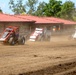 A dirt track racing event featuring multiple open-wheel sprint cars or midget race cars competing on a dusty oval circuit. At least three vehicles are visible racing in close proximity, kicking up significant dust clouds. The race appears to be taking place at a fairgrounds-style venue with a red-roofed building and trees visible in the background.